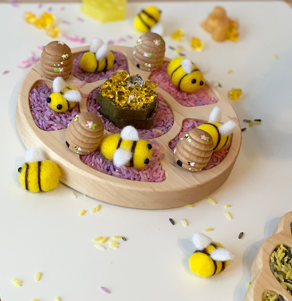 Round wooden Tinker Tray with purple rice, felt bees, wooden beehives, and yellow gem on white surface