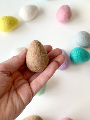 Hand holding a single brown felt Easter egg with pastel felt eggs scattered on white surface background