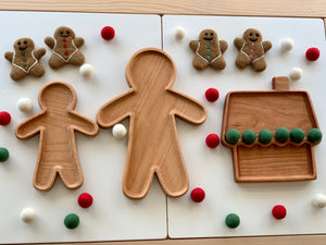 Large wooden person-shaped and house-shaped sensory trays surrounded by gingerbread figures and red, white, green felt balls on white surface