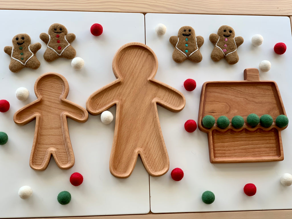 Large wooden person-shaped and house-shaped sensory trays surrounded by gingerbread figures and red, white, green felt balls on white surface