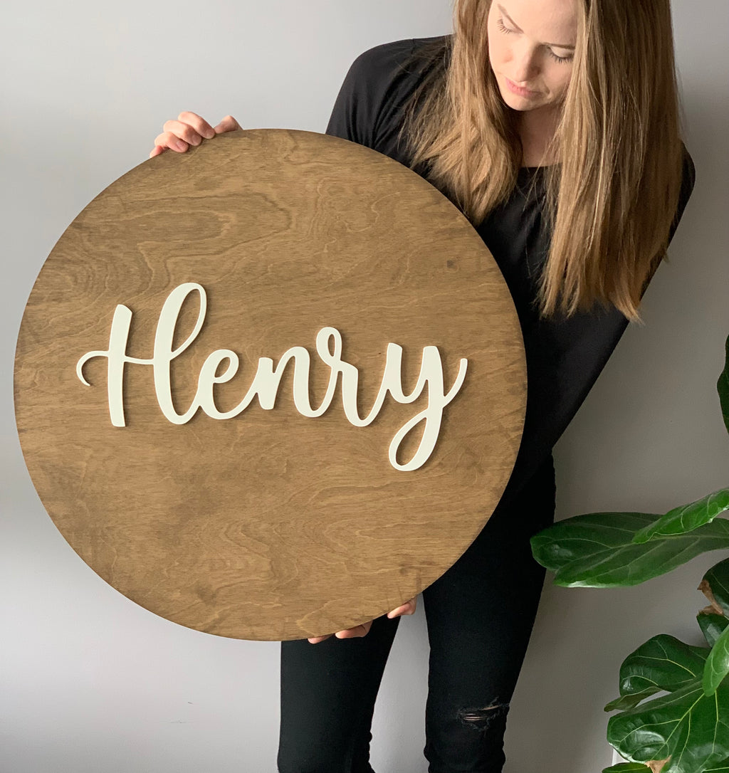Woman holding large round wooden name sign with white cursive Henry lettering on natural wood background