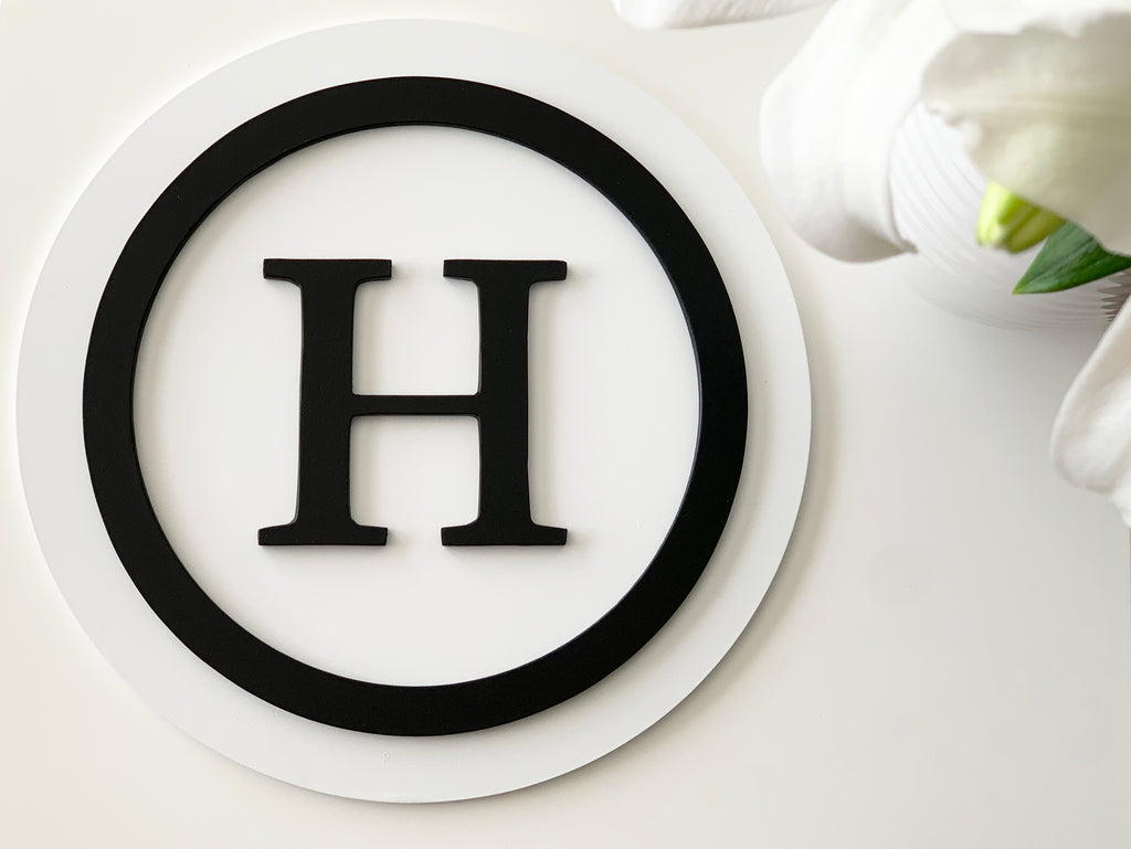 Round 3D monogram sign with black letter H and circle on layered white wooden background with flowers nearby