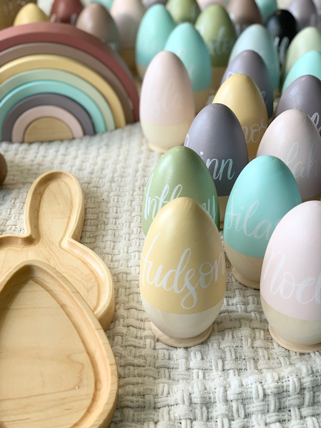 Assorted pastel hollow wooden Easter eggs with white cursive names displayed on cream textured fabric with wooden bunny tray and rainbow toy