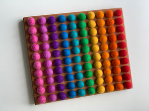 Wooden sensory tray with smooth engraved holes holding colorful felt balls arranged in rainbow rows on white background