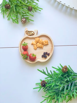 Natural wooden apple-shaped sensory tray with animal crackers, strawberries, nuts, and dried berries on white surface with pine branches