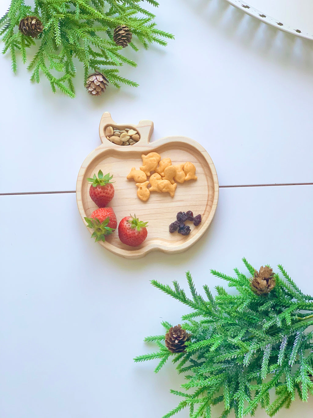Natural wooden apple-shaped sensory tray with animal crackers, strawberries, nuts, and dried berries on white surface with pine branches