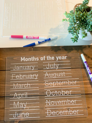 Clear acrylic dry erase board with dotted month names for tracing and three colored markers on wooden table with plant nearby