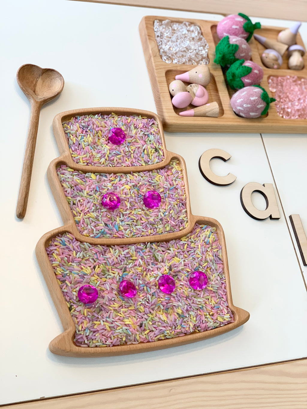 Wooden cake-shaped sensory tray filled with colorful rice and pink gem decorations with a wooden heart spoon nearby