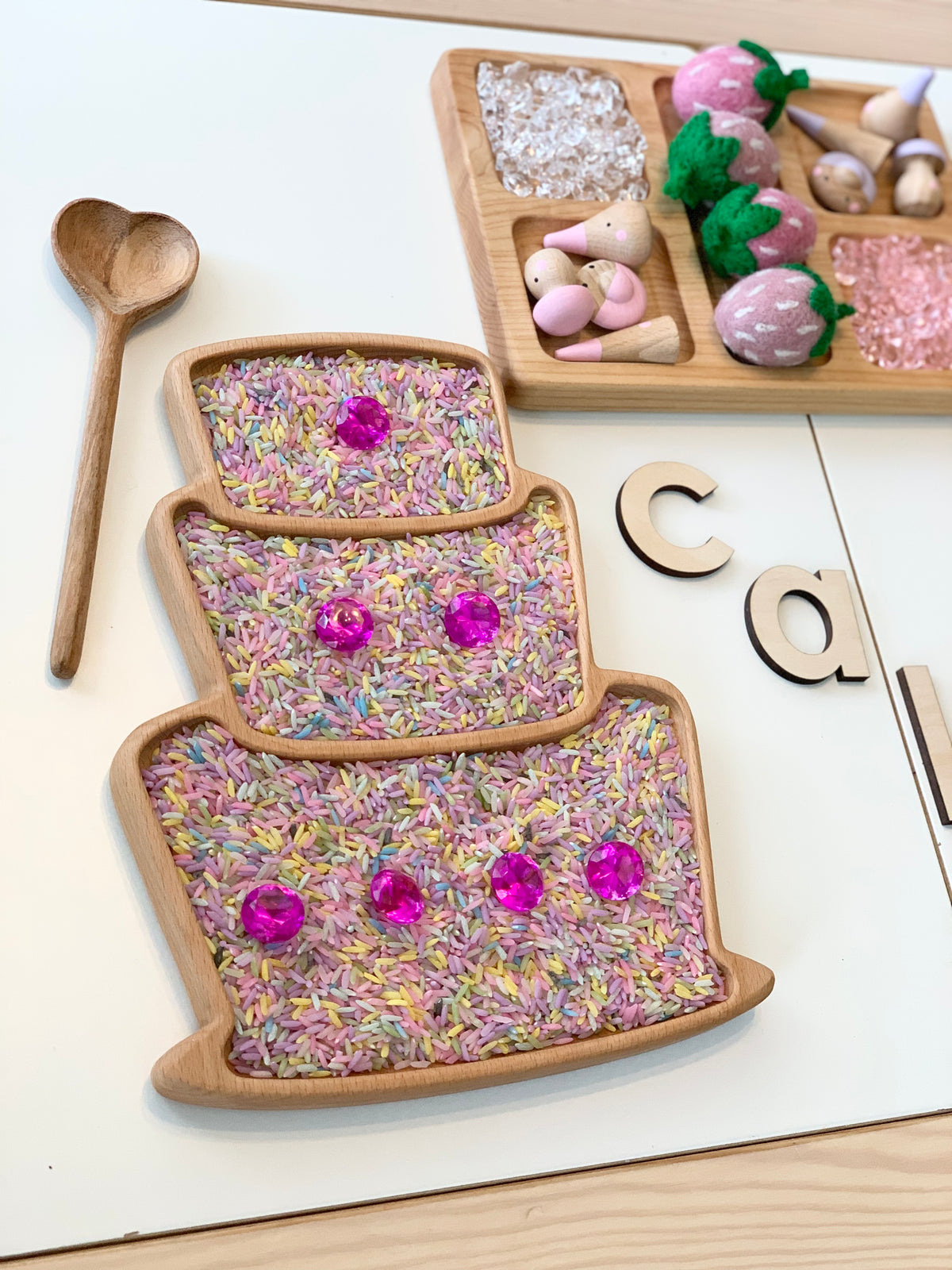 Wooden cake-shaped sensory tray filled with colorful rice and pink gem decorations with a wooden heart spoon nearby