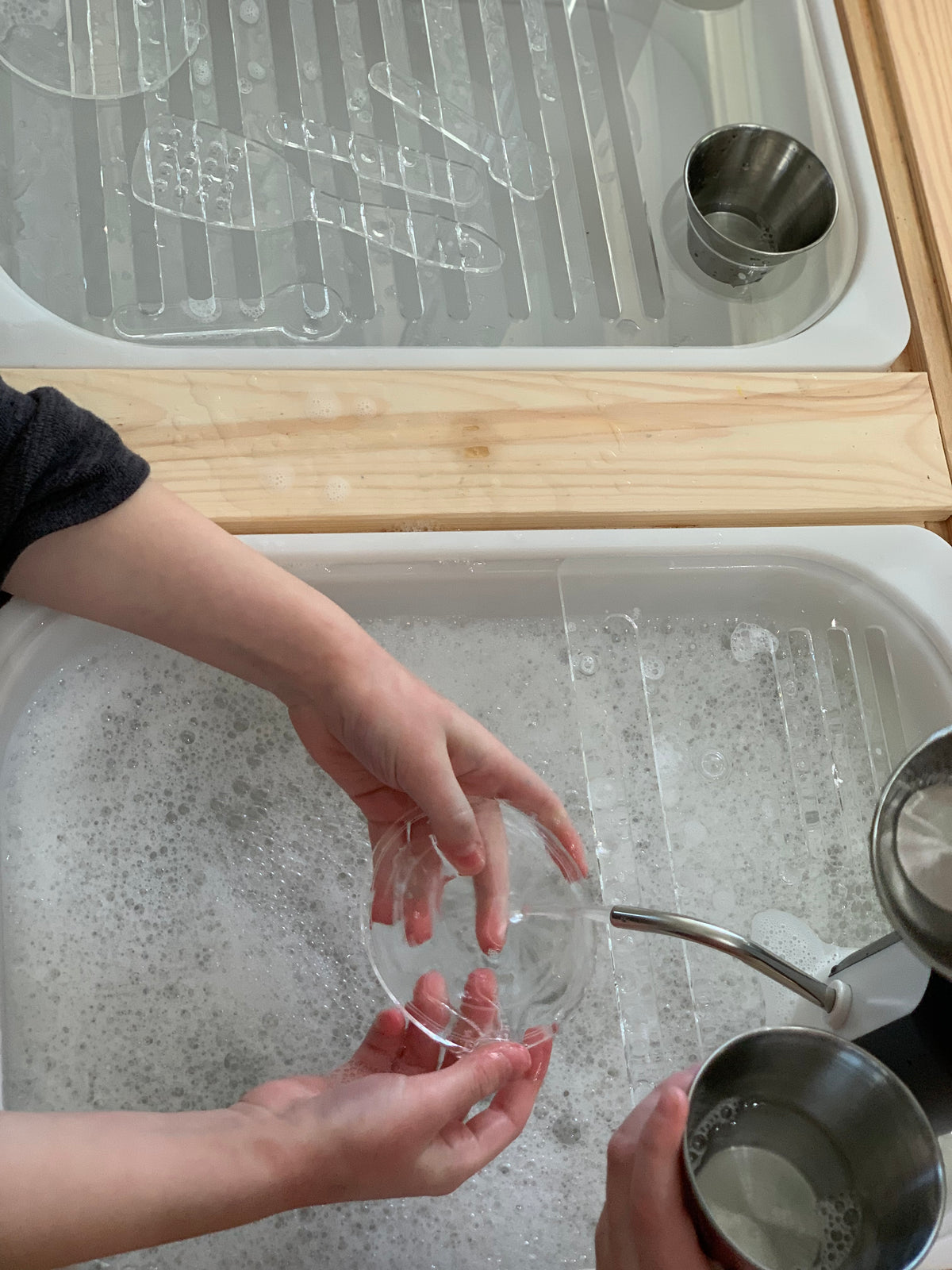 Child washing clear plastic cup in white play kitchen sink insert with water and soap bubbles on wooden surface
