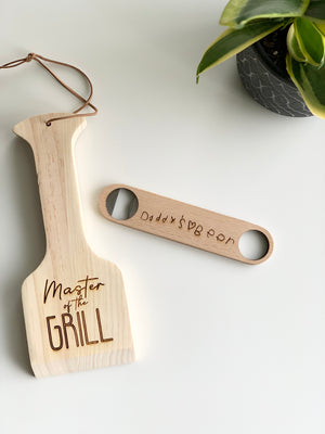 Custom wooden barbecue scraper engraved with "Master of the Grill" and wooden bottle opener with handwritten engraving on white surface