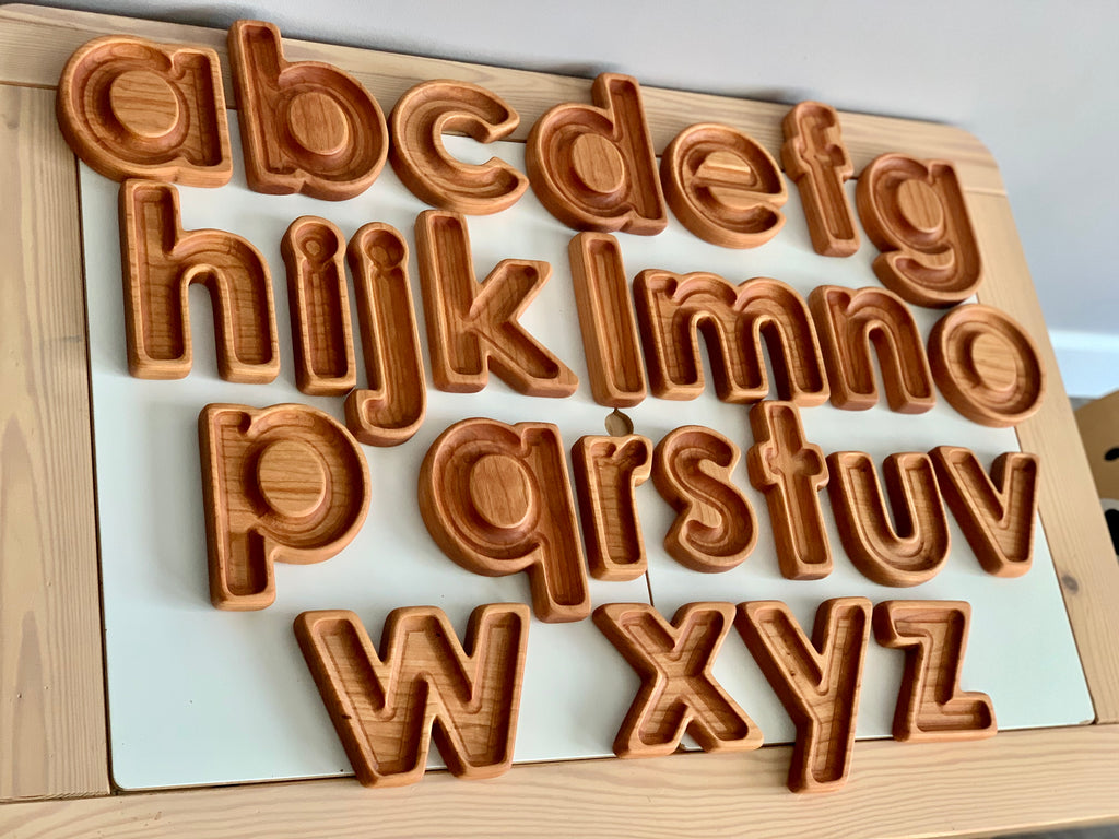 Natural wooden lowercase alphabet sensory trays arranged on white board with light wood frame