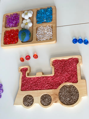 Natural wooden train-shaped sensory tray filled with colored rice and a wooden tray with colorful sensory beads on white surface