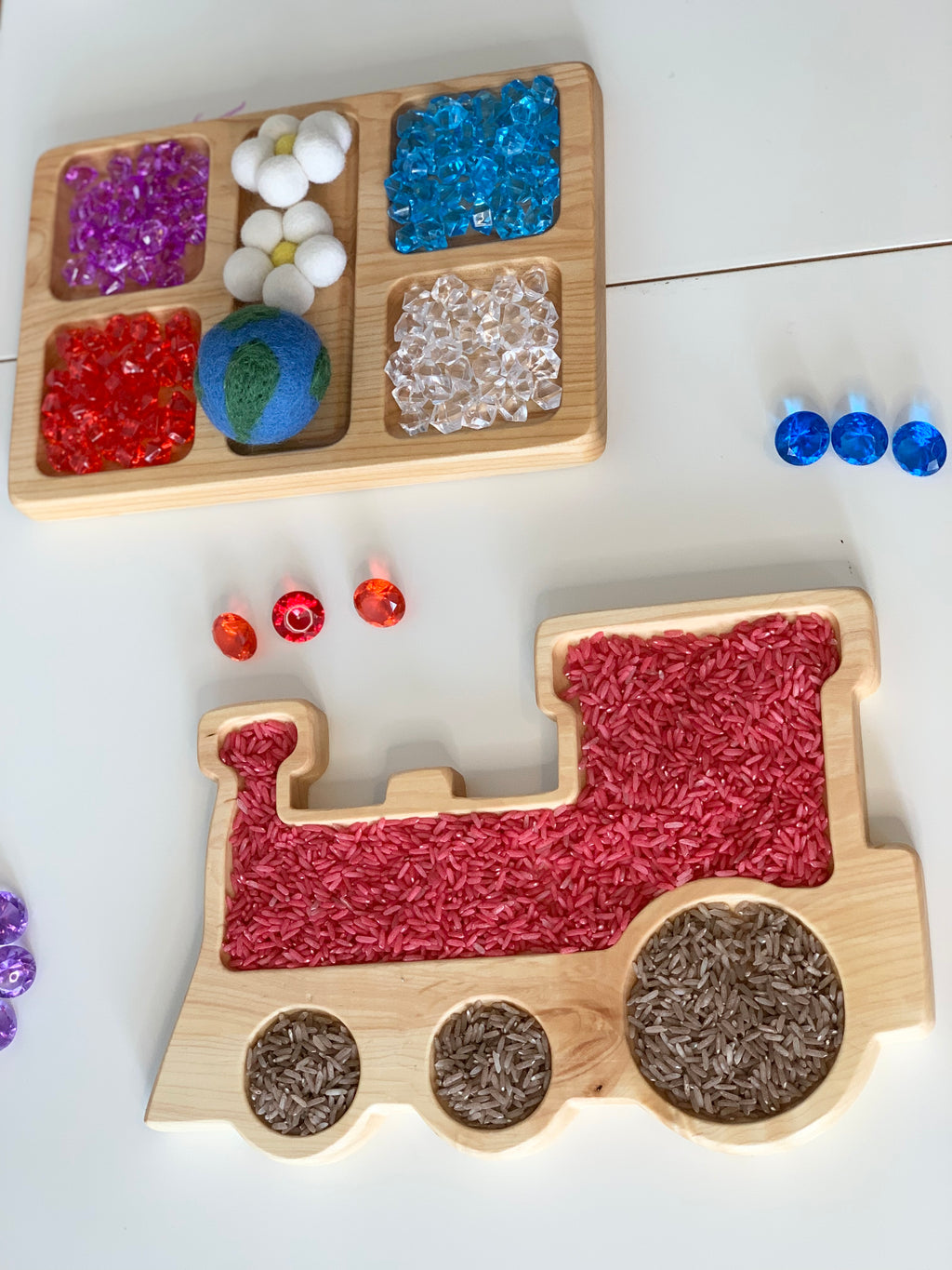 Natural wooden train-shaped sensory tray filled with colored rice and a wooden tray with colorful sensory beads on white surface