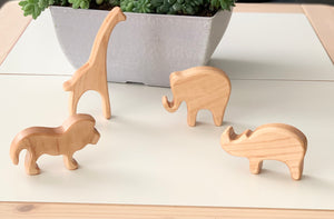 Natural wooden safari animal toy figurines including lion, giraffe, elephant, and rhino on white surface with plant pot background