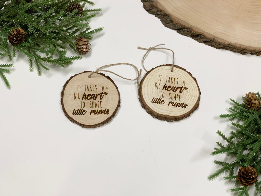Two round wood burned Christmas ornaments with "It takes a big heart to shape little minds" text on white background with pine branches