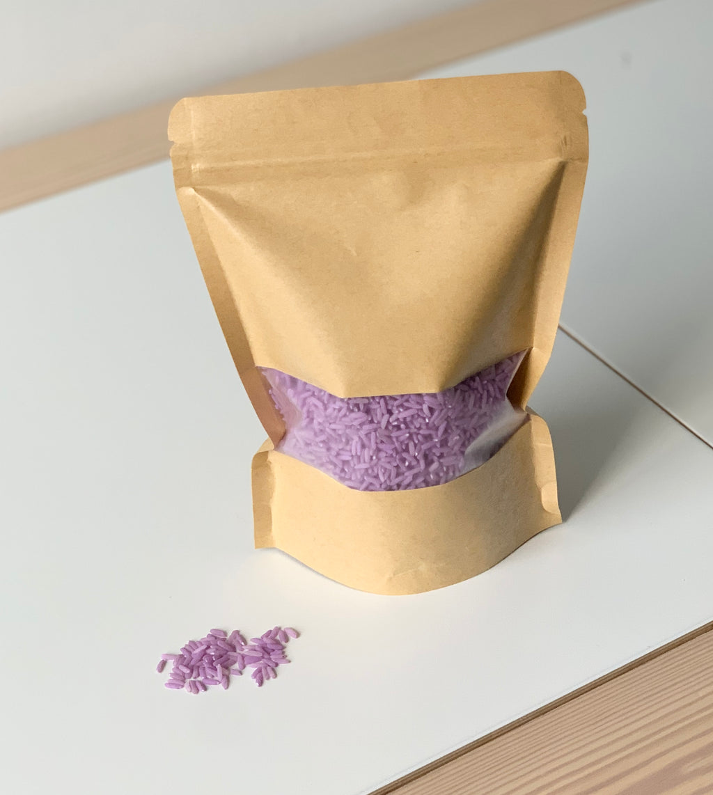 Brown resealable paper pouch partially filled with purple colored sensory rice spill on white surface