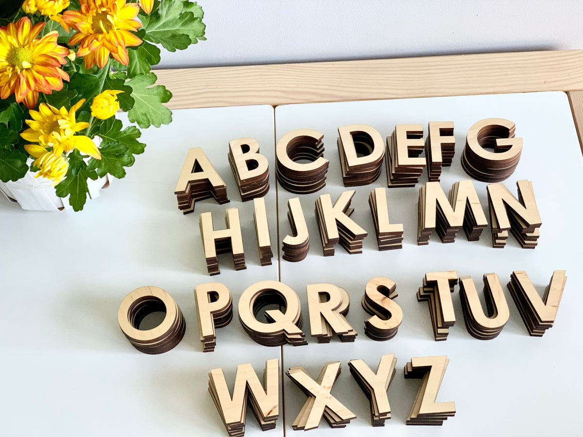 Natural wooden uppercase alphabet letters in stacks arranged on white surface next to yellow and orange flowers