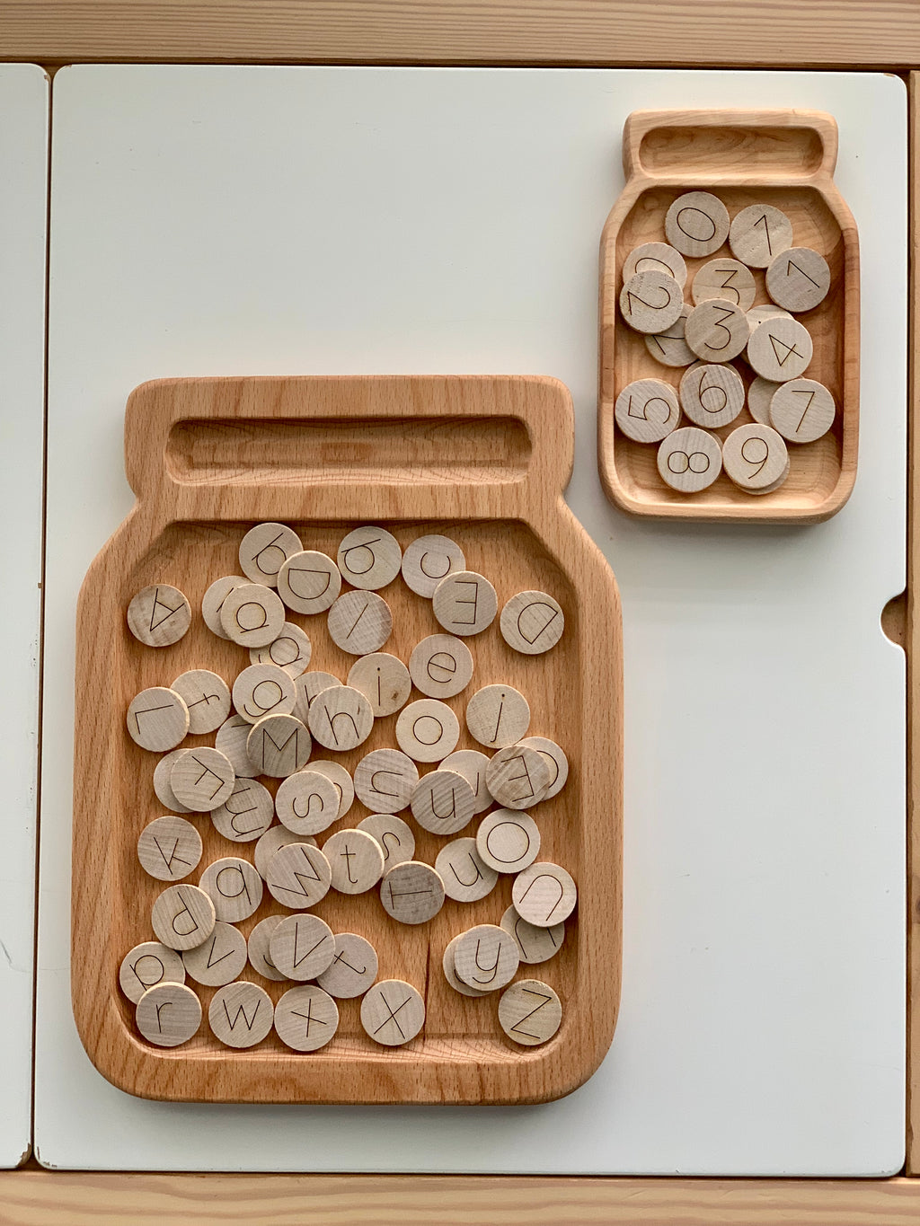 Set of natural wooden 1-inch round alphabet and number discs with simple line engraving in jar-shaped trays on white table