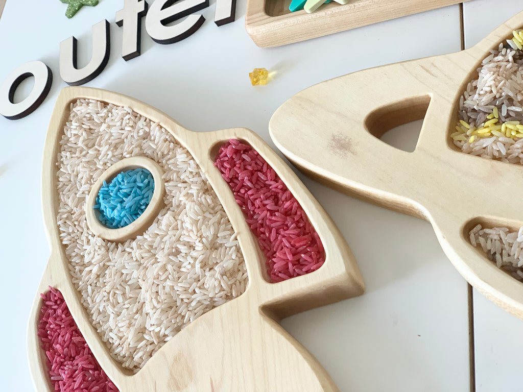 Natural wooden rocket ship sensory tray filled with colored and white rice on white surface with letters