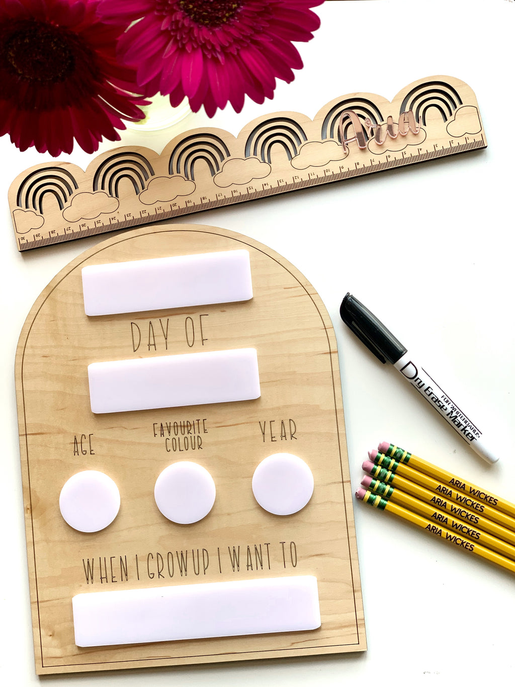 Wooden dry erase school sign with writeable sections, black marker, personalized pencils, rainbow ruler, and pink flowers on white background