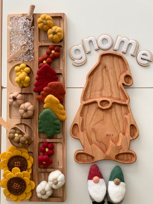Natural wooden gnome-shaped sensory tray beside wooden tray with colorful felt flowers and ice-like beads on white surface