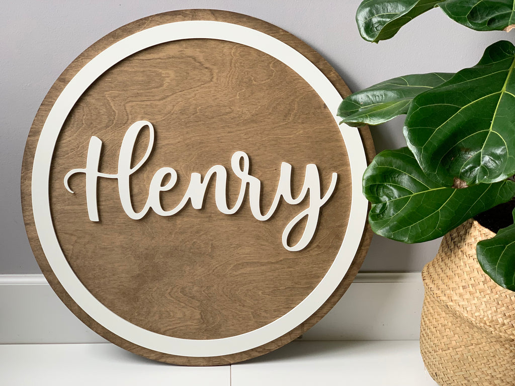 Round wooden name sign with white cursive letters and circular border on natural wood background next to green plant