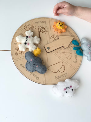 Round wooden weather wheel with engraved weather icons and felt weather character toys on white surface with hand
