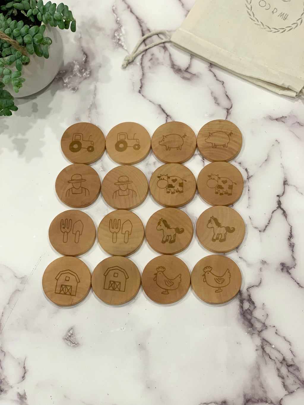 Natural wooden round memory game pieces engraved with farm animals, tractor, farmer, barn, and utensils on marble surface near plant and pouch