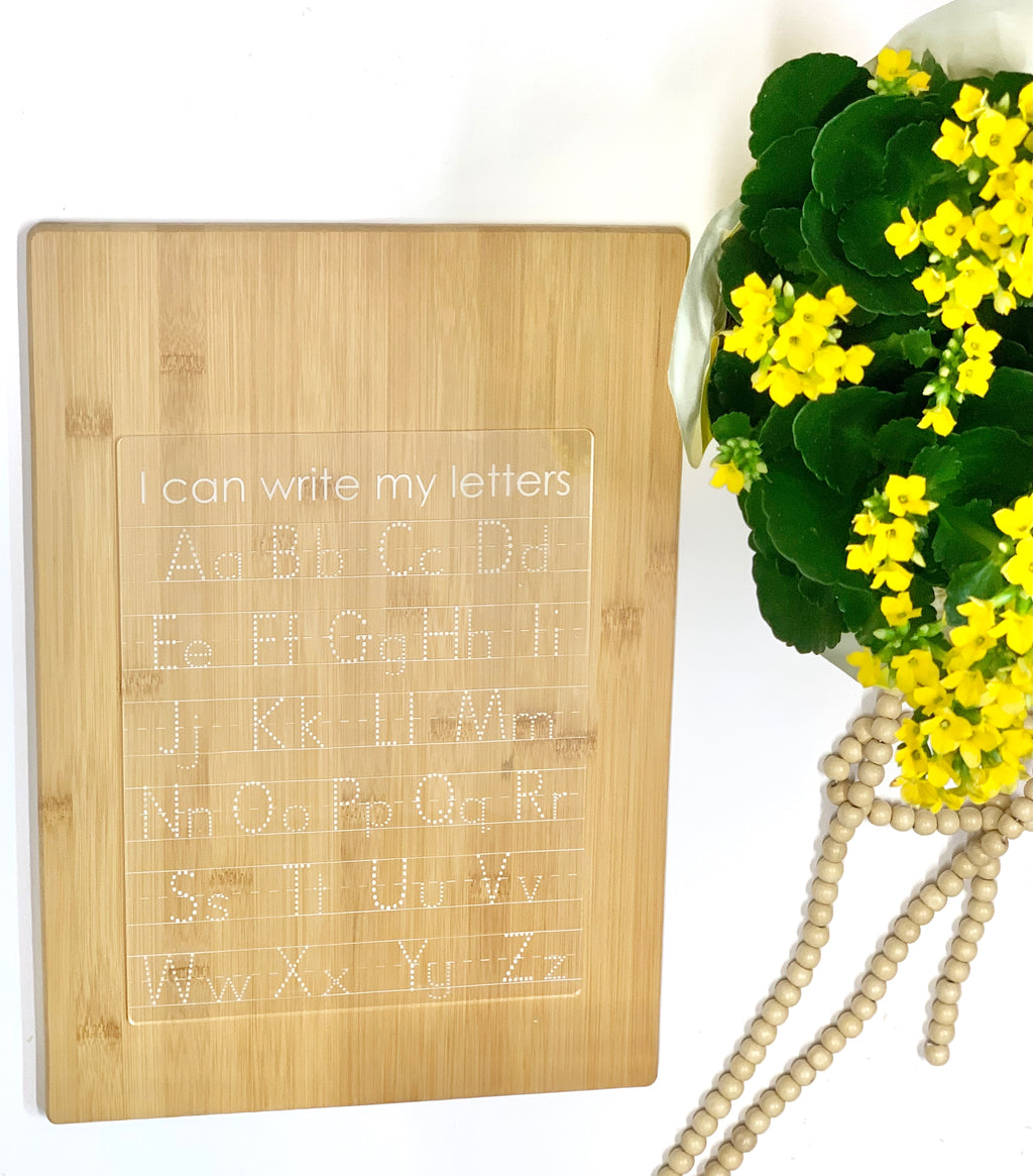 Acrylic dry erase tracing board with uppercase and lowercase letters engraved on natural wood background next to yellow flowers