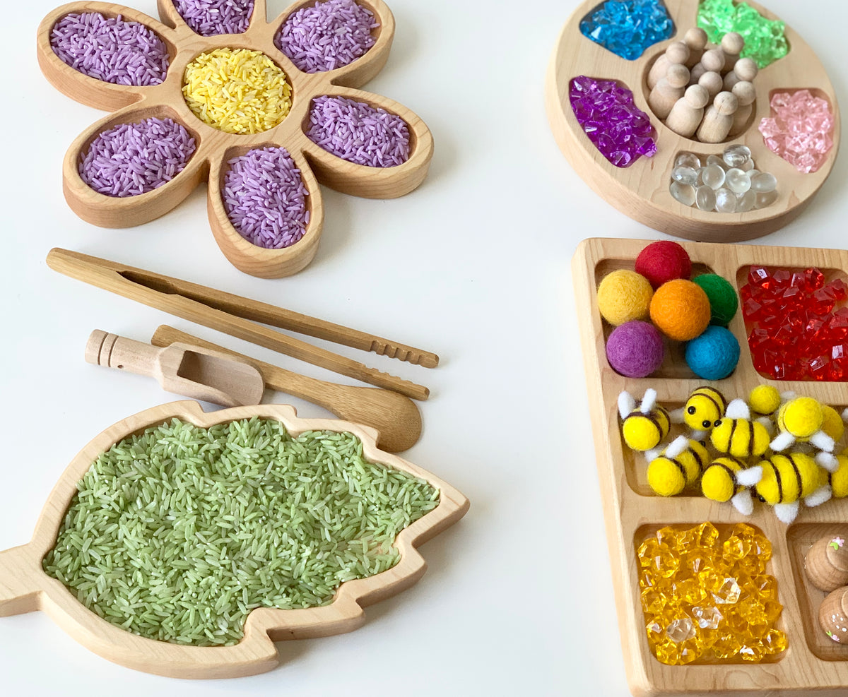 Natural birch wood leaf-shaped sensory tray filled with green rice and wooden scoop and tongs on white background