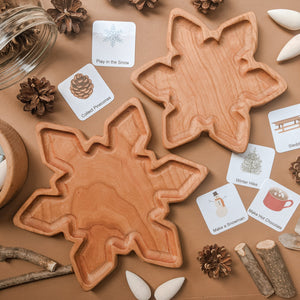 Two natural wooden snowflake-shaped trays on brown surface surrounded by pinecones, cards, and small wood pieces