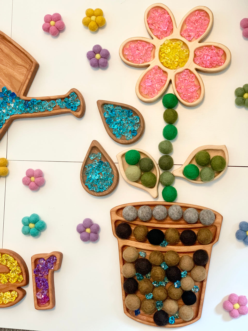 Flower pot and watering can wooden sensory trays filled with colored beads and felt balls on white surface