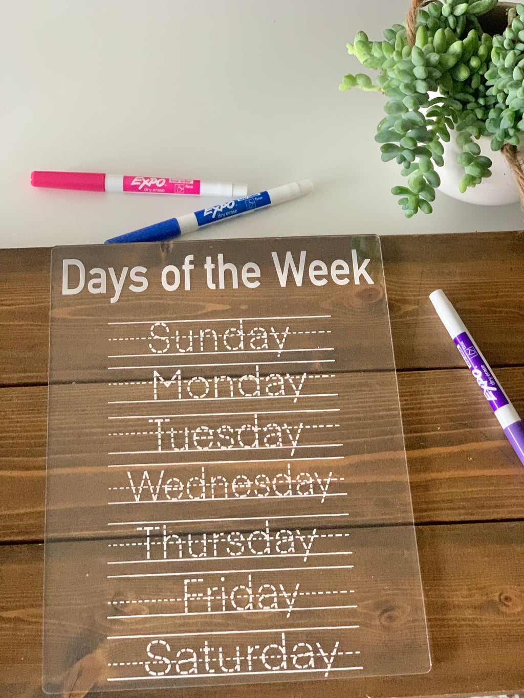 Clear acrylic dry erase board with days of the week tracing letters and colorful dry erase markers on wood table