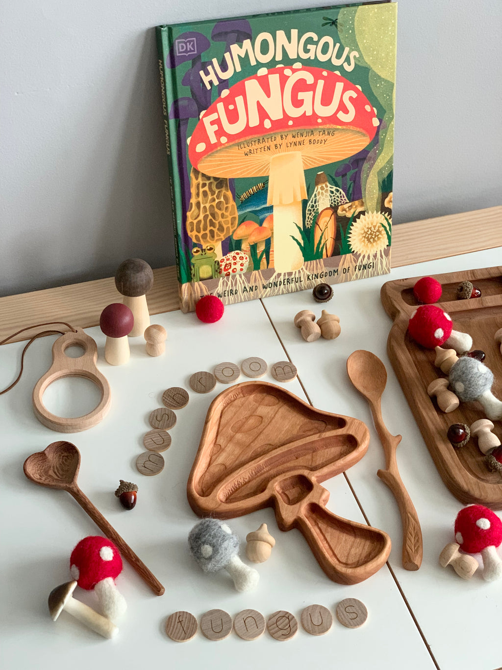 Wooden mushroom-shaped sensory tray surrounded by natural wooden utensils and mushroom toys with fungus book background