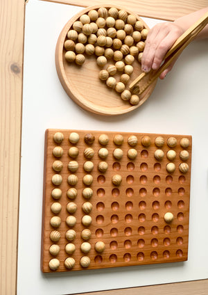 Wooden 100 frame sorting board with engraved holes and natural wood balls with child's hand using wooden tongs on white background