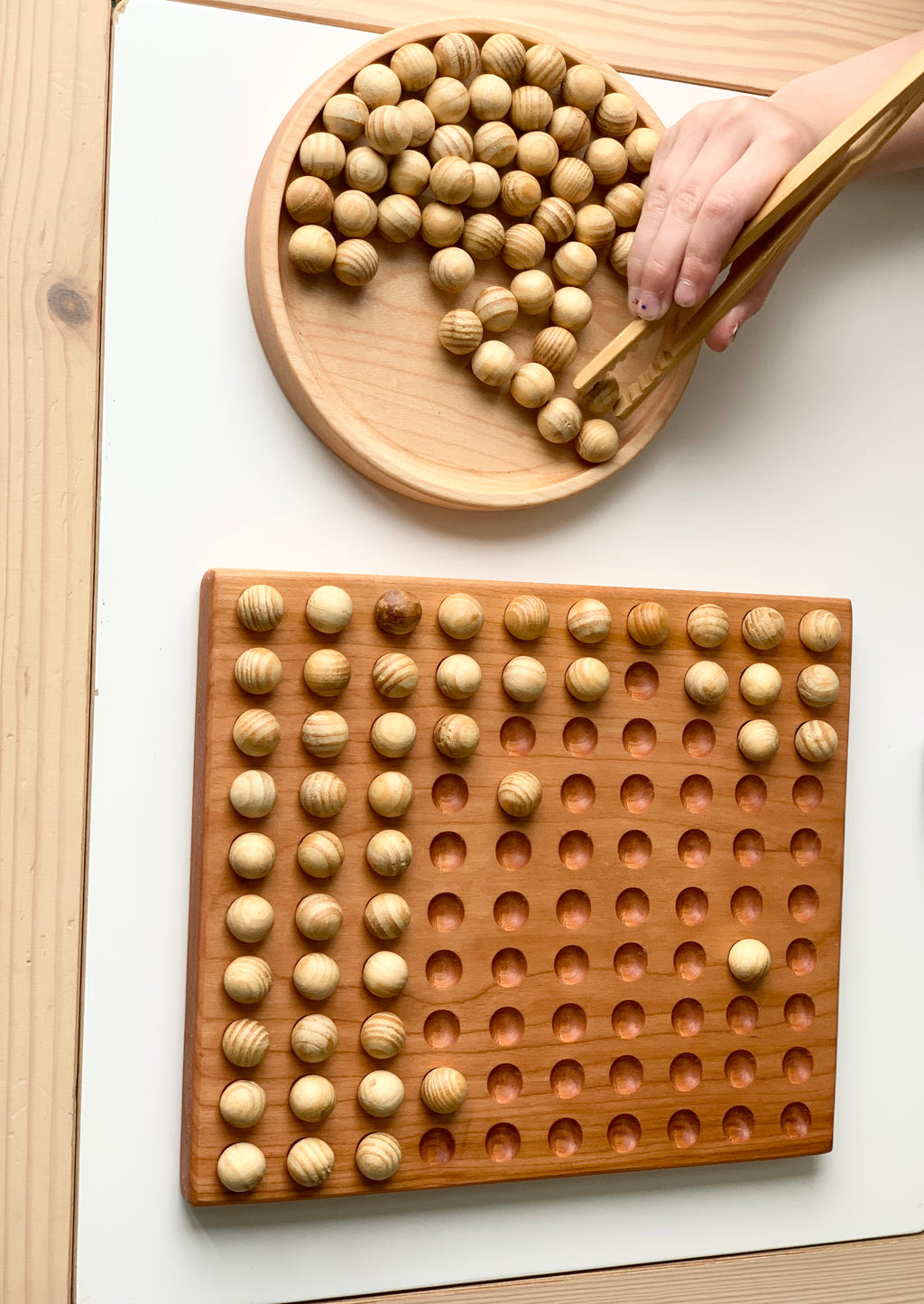 Wooden 100 frame sorting board with engraved holes and natural wood balls with child's hand using wooden tongs on white background
