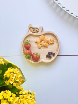 Natural wooden apple-shaped sensory tray with strawberries, goldfish crackers, raisins, and pumpkin seeds on white table
