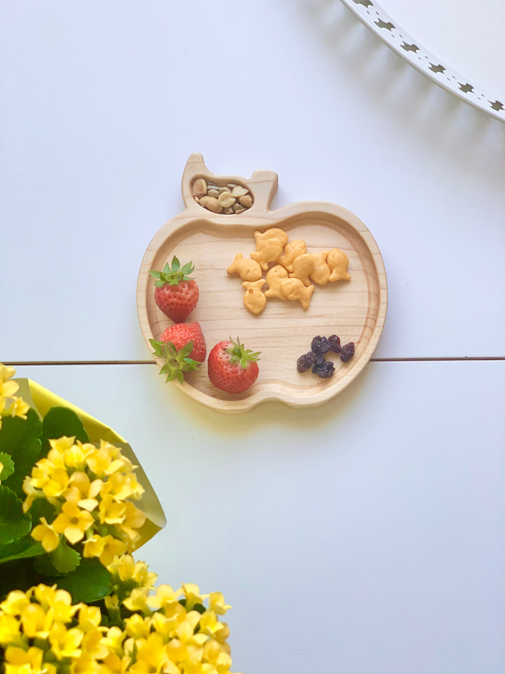 Natural wooden apple-shaped sensory tray with strawberries, goldfish crackers, raisins, and pumpkin seeds on white table