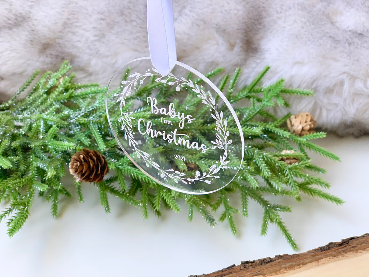 Clear acrylic baby’s first Christmas ornament with white engraved lettering and floral wreath design hanging on greenery background