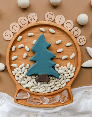 Round wooden sensory tray shaped like a snow globe with blue tree, white beans, and clear crystals on brown background