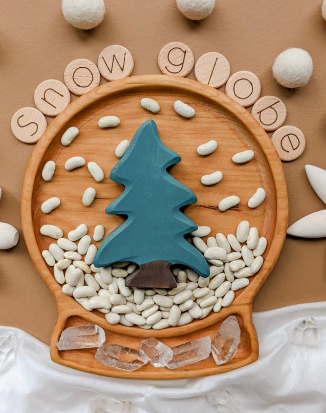 Round wooden sensory tray shaped like a snow globe with blue tree, white beans, and clear crystals on brown background