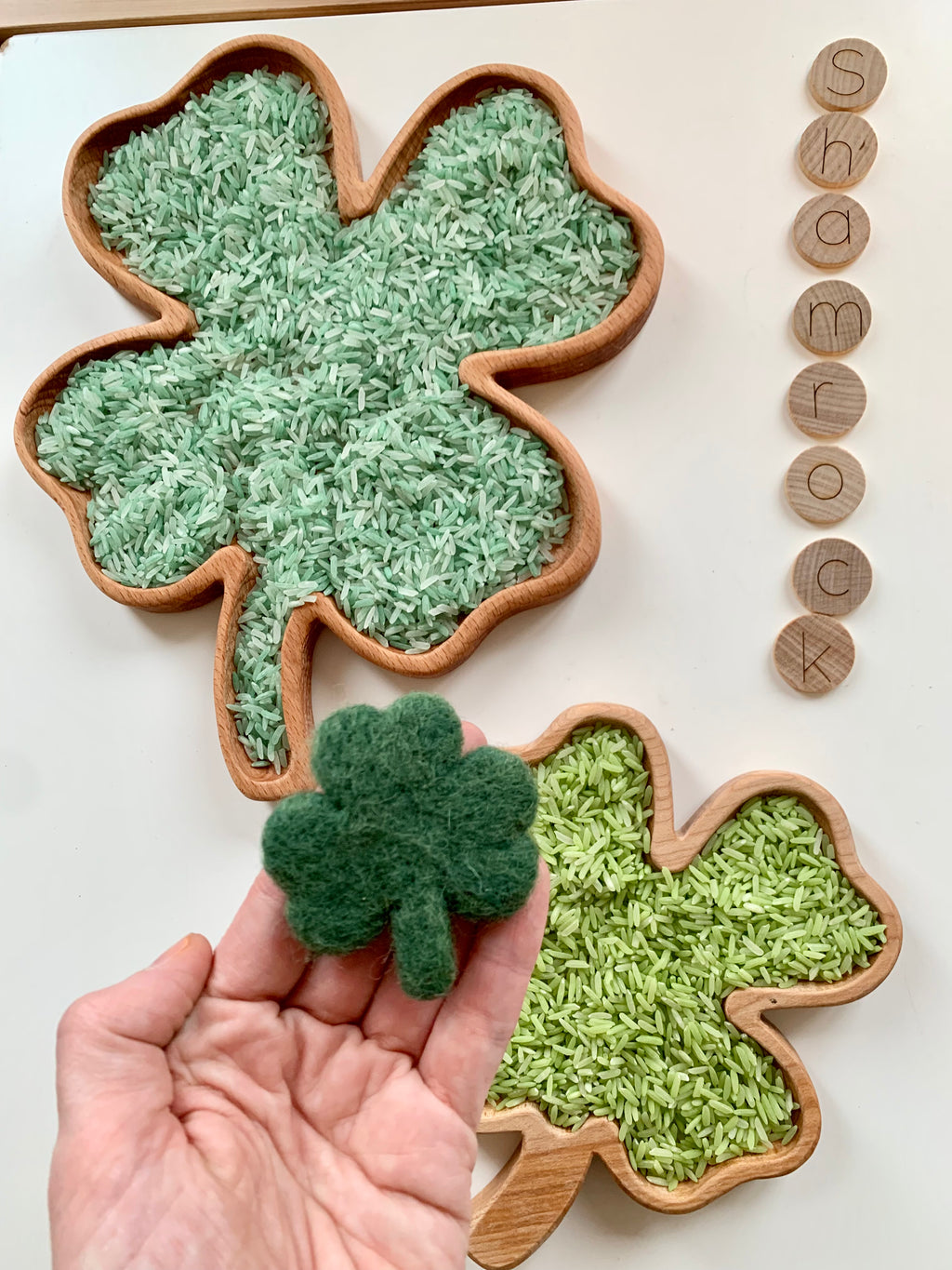 Wooden shamrock-shaped sensory trays filled with green rice and hand holding a small dark green felt clover toy