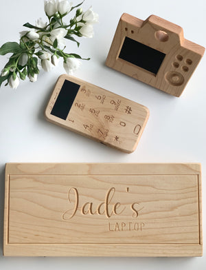 Natural wooden toy camera and phone with engraved details and black screen alongside engraved wooden laptop toy on white background with flowers