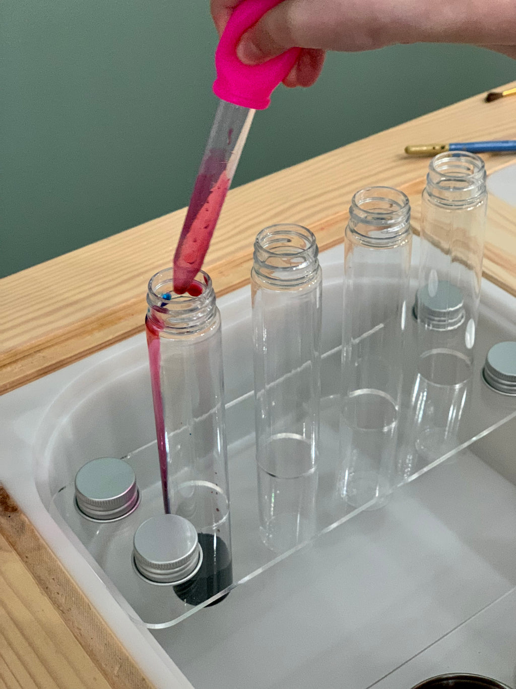 Clear acrylic Flisat insert holding four test tubes with metal lids and child using pink dropper to fill one tube