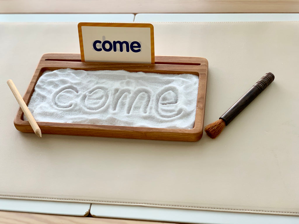 Rectangular wooden sensory tray filled with white sand, word "come" drawn, with flash card and wooden stylus on cream surface
