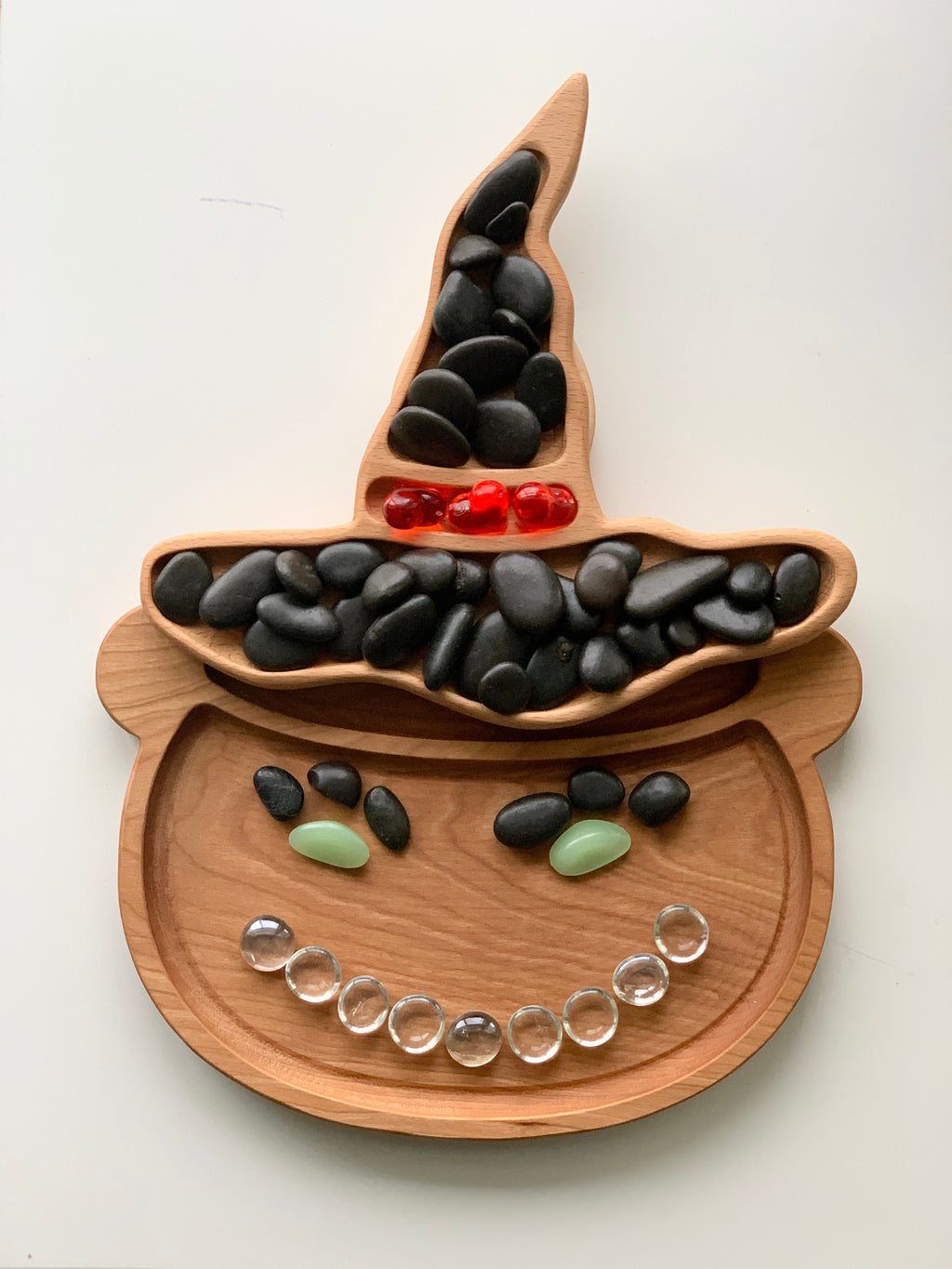 Natural wooden witch hat shaped sensory tray with black, red, green, and clear glass stones arranged in face pattern