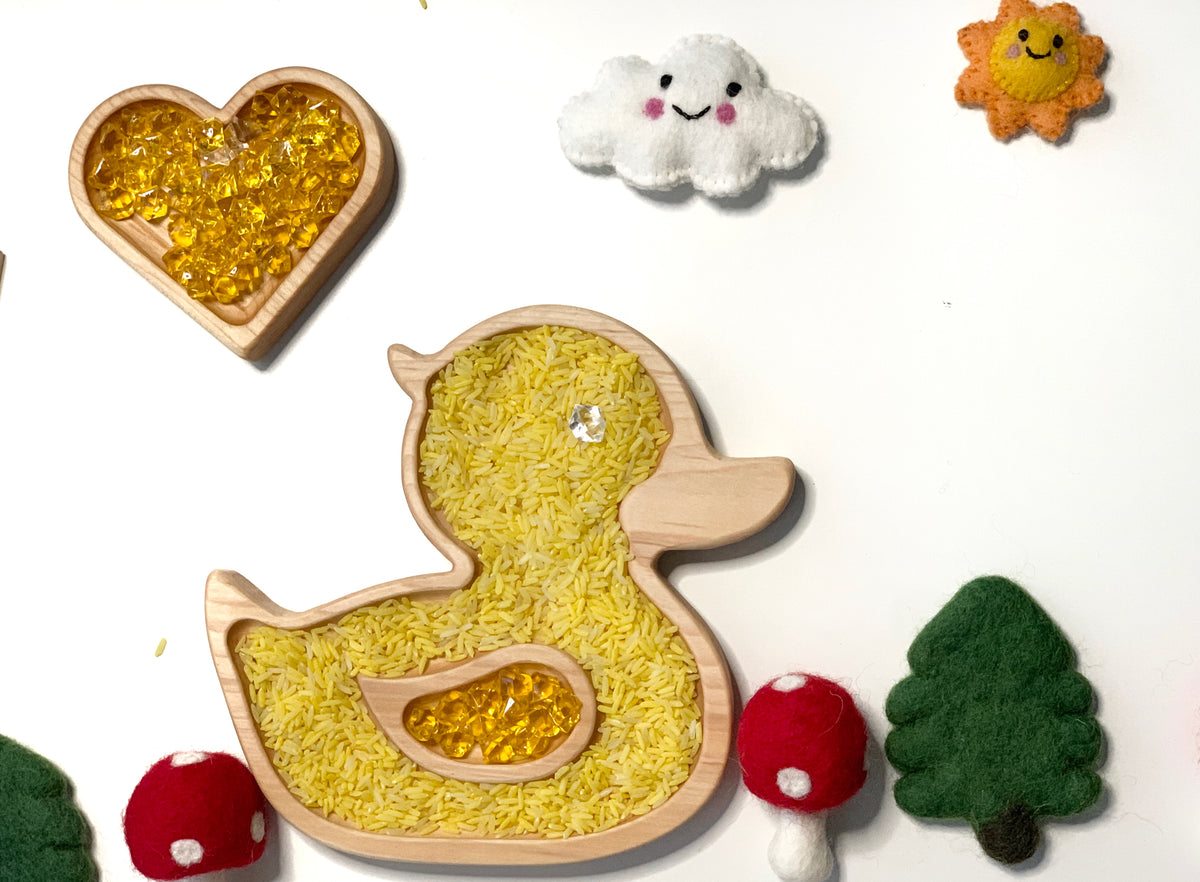 Wooden duck-shaped sensory tray filled with yellow rice and amber gems surrounded by felt clouds, sun, trees, and mushrooms