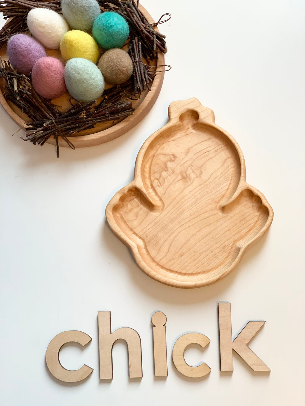 Wooden chick-shaped sensory tray with colorful felt eggs in twig nest and wooden letters spelling chick on white surface