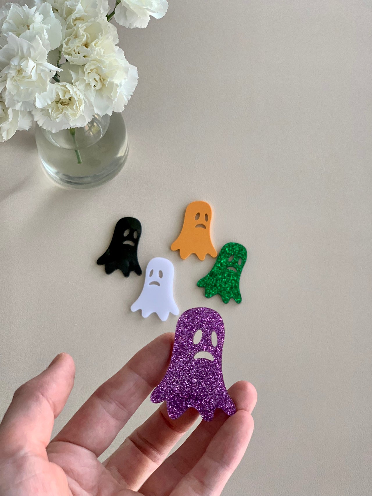 Set of five laser cut acrylic ghost shapes in purple glitter, white, black, orange, and green on beige surface with flowers nearby